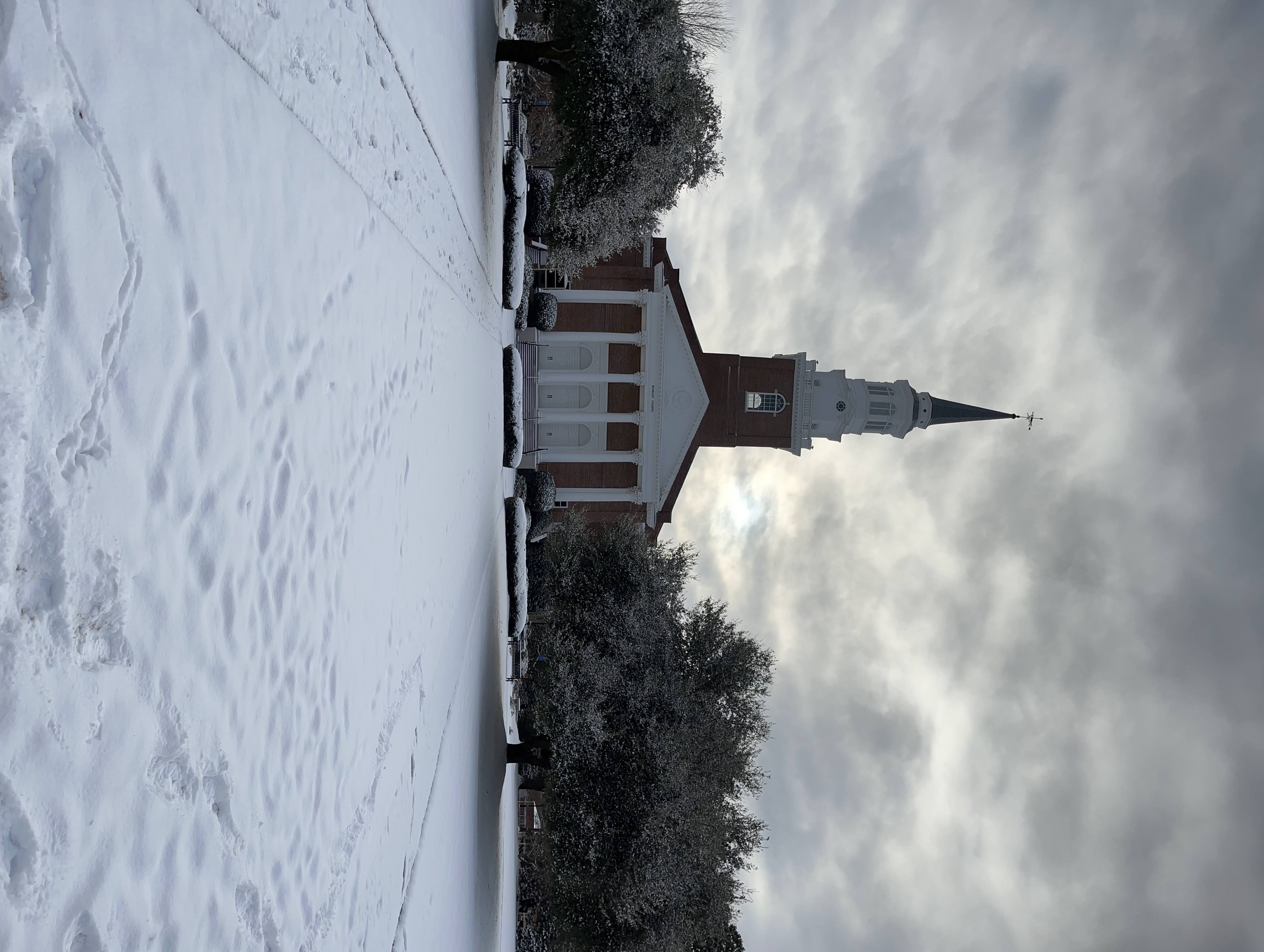 The steeple and brick facade of a small-town chapel under a heavy sky, with fresh snow across the grounds.