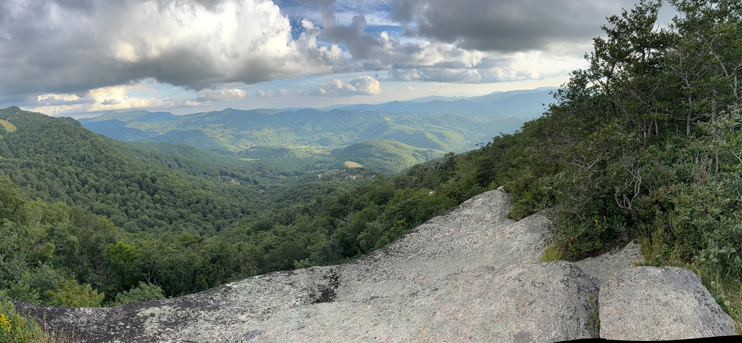 A wide summer panorama of layered Blue Ridge mountains beneath dramatic clouds from Beech Mountain.