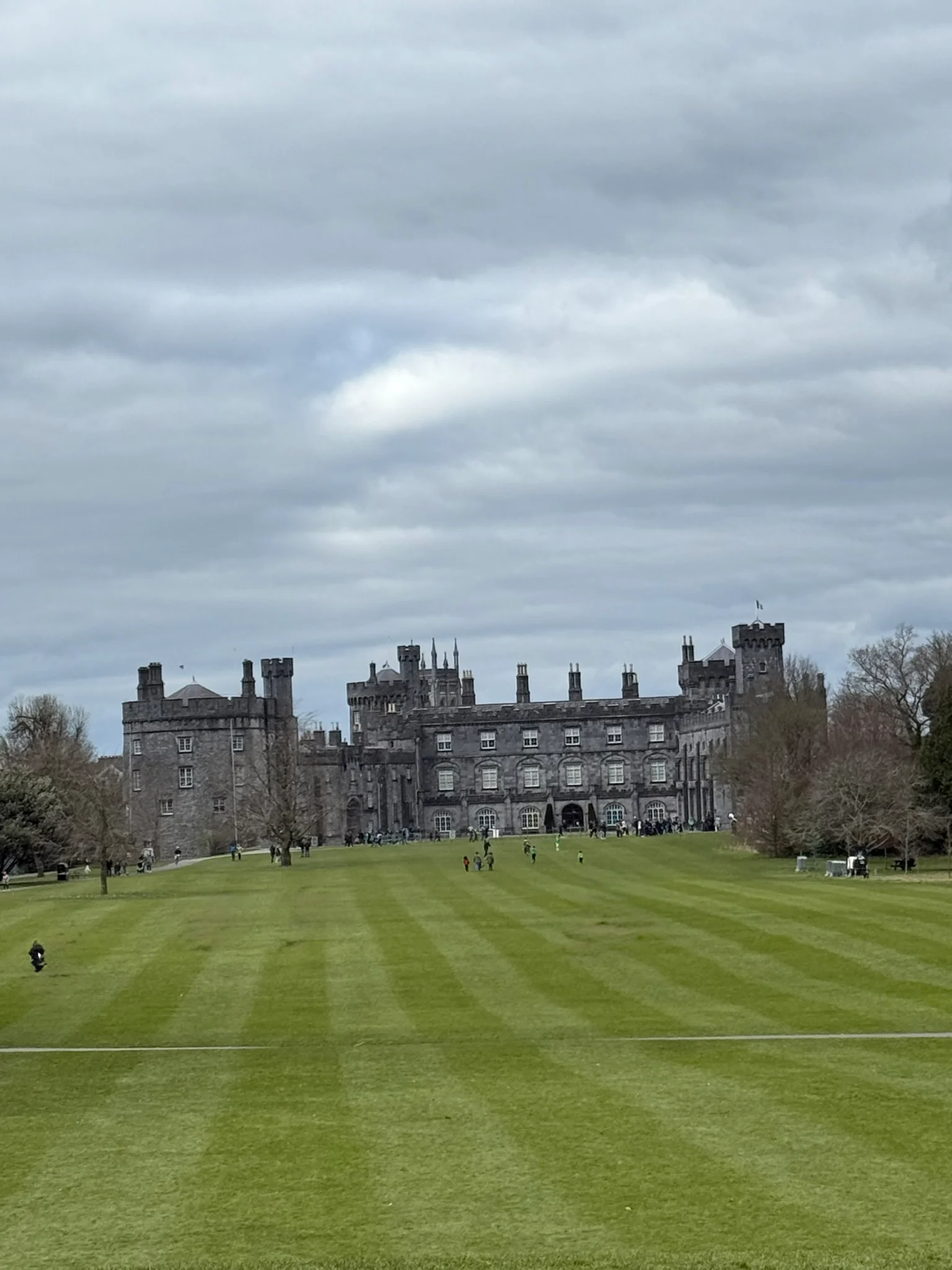 Kilkenny Castle seen across a wide green lawn under a cloudy March sky.