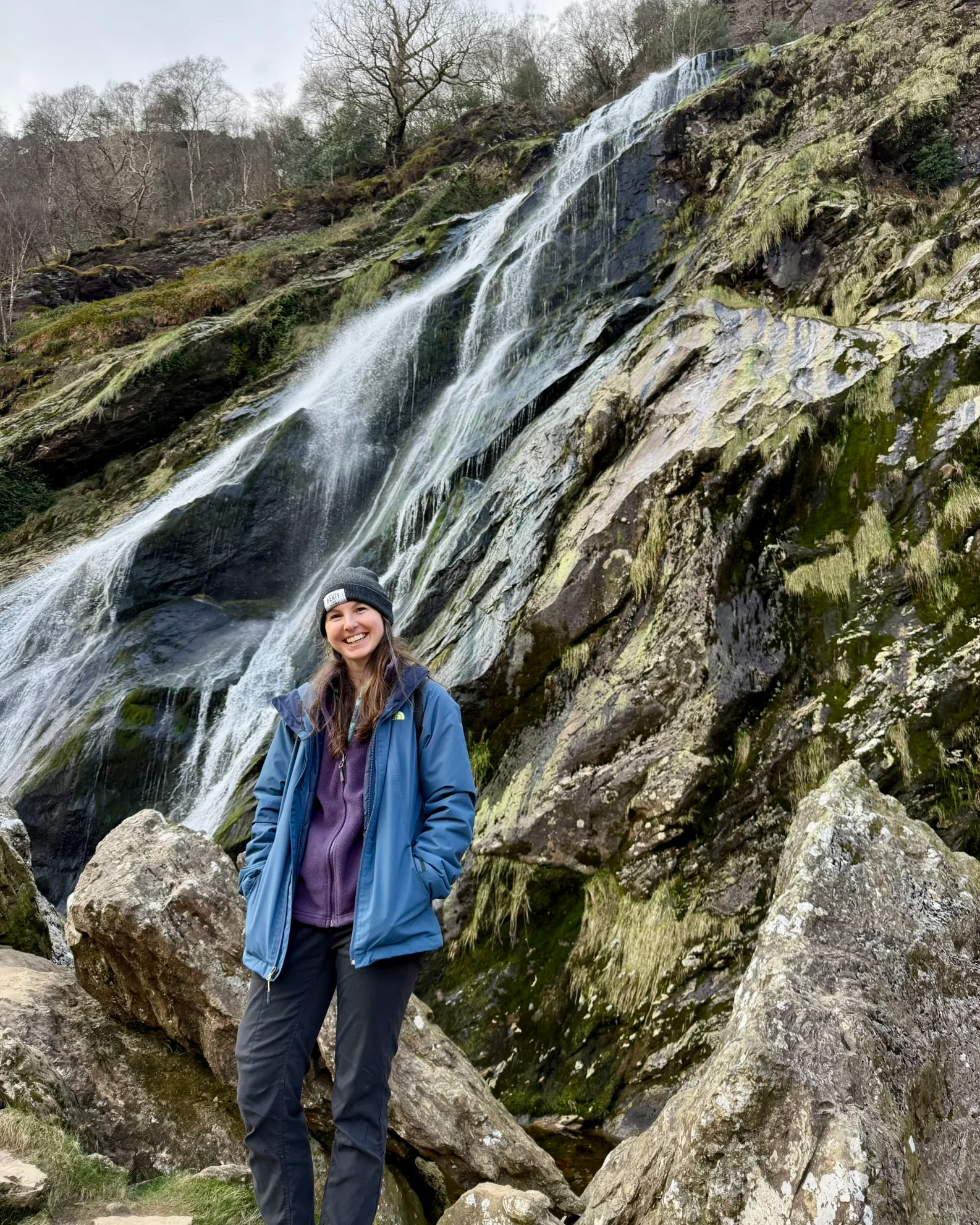 Sydney standing beside Powerscourt Waterfall as water runs over mossy Wicklow rocks.